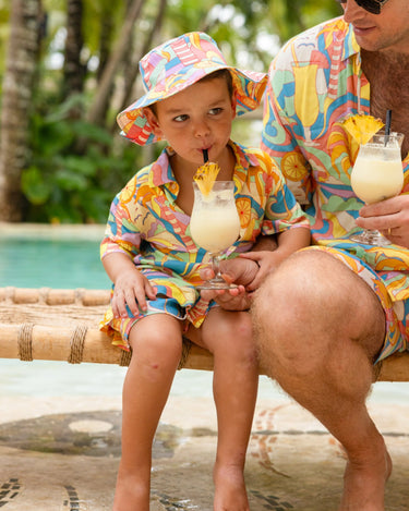 Man and child in matching colorful outfits sitting by a pool with drinks.