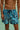 A man wearing blue and black abstract pattern swim trunks standing in shallow turquoise pool water.