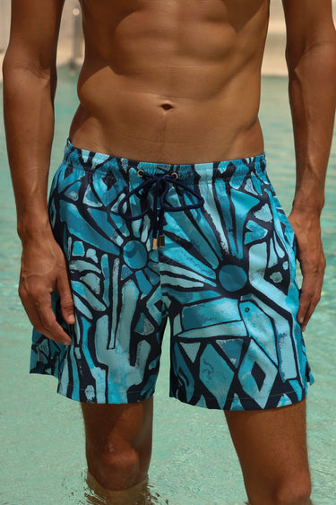 A man wearing blue and black abstract pattern swim trunks standing in shallow turquoise pool water.