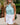 Woman wearing a light blue polo shirt and white skirt standing outdoors with greenery in the background