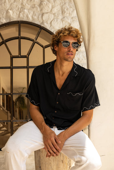 A man wearing a black short-sleeve button-up shirt with white wavy contrast stitching and white pants, sitting outdoors in front of a textured white wall and arched window.