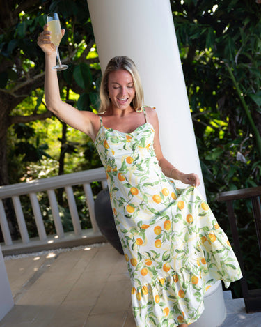 Woman in a floral dress holding a glass, standing on a patio with greenery in the background